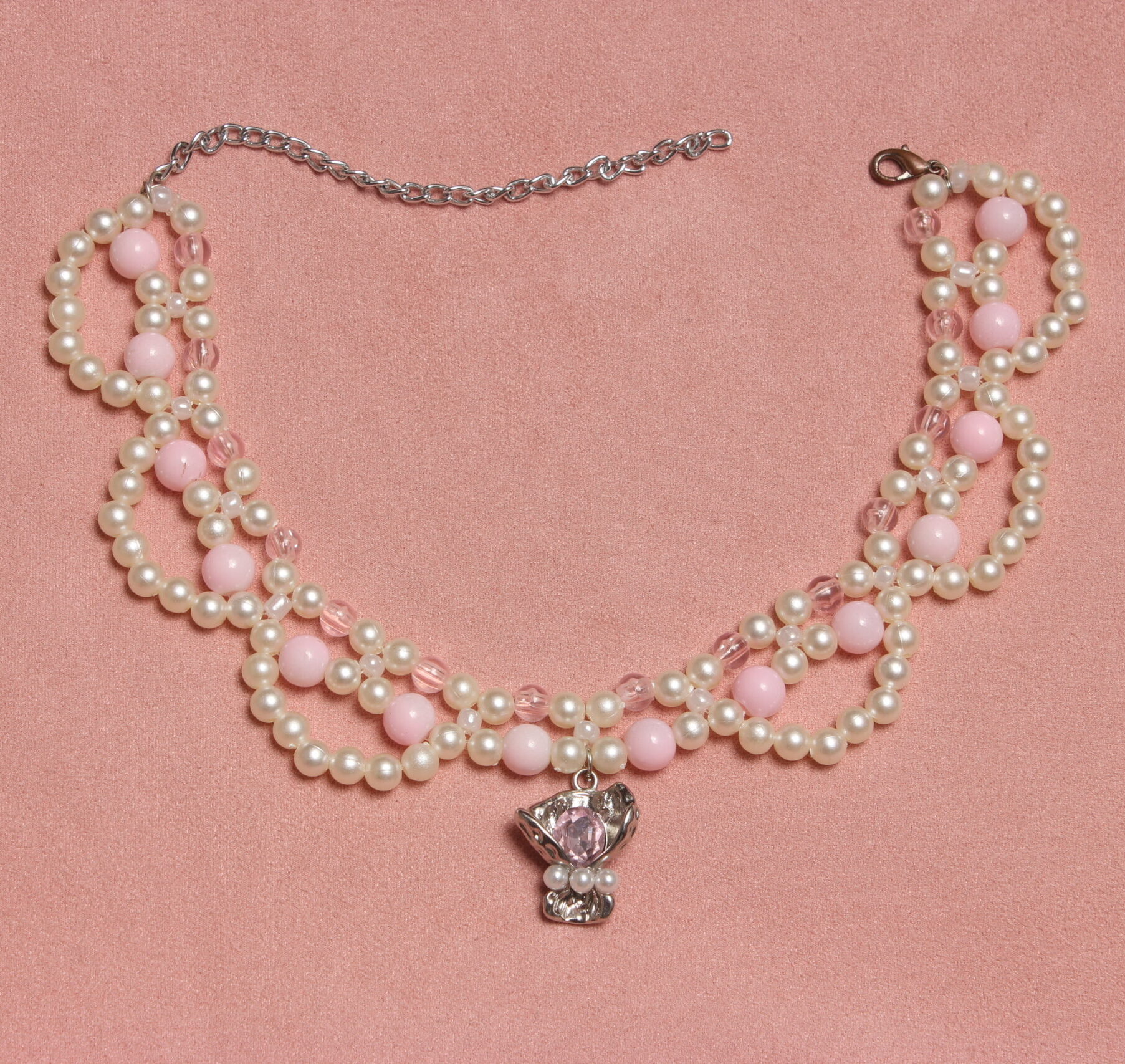 Collier en perles vintage. 2 rangées de perles blanches et rose pales et une troisième rangée formant des arcs de cercles en perles blanches. Pendentif argenté avec une pierre rose pâle et 3 petites perles blanches nacrées au centre du collier. Taille ajustable grâce à la chaînette argentée.