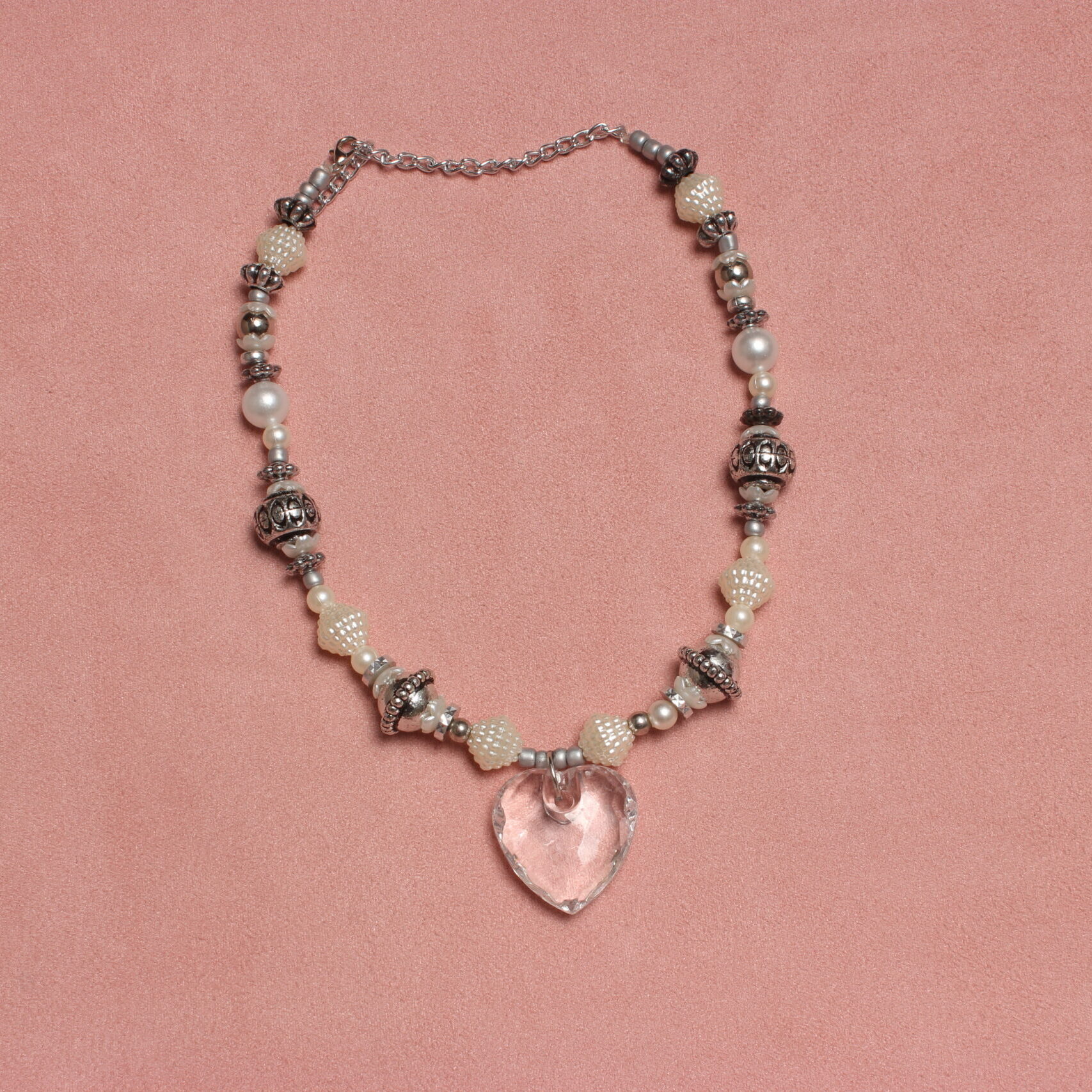 Collier en perles vintage. Alternance de perles blanches nacrées et grises. Pendentif en forme de cœur transparent au centre. Formes et grosseurs variées. Taille ajustable grâce à la chaînette argentée