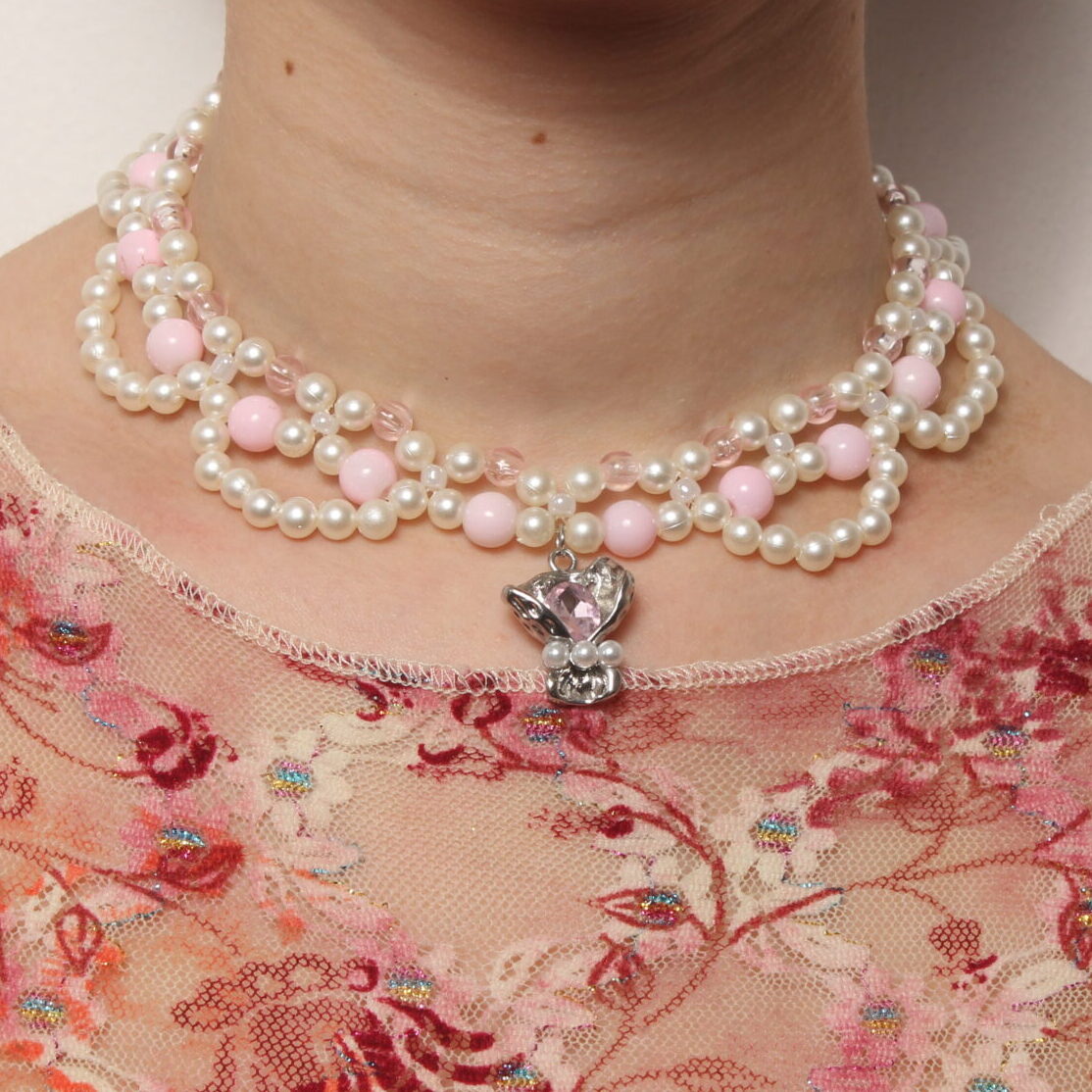 Collier en perles vintage. 2 rangées de perles blanches et rose pales et une troisième rangée formant des arcs de cercles en perles blanches. Pendentif argenté avec une pierre rose pâle et 3 petites perles blanches nacrées au centre du collier. Taille ajustable grâce à la chaînette argentée.