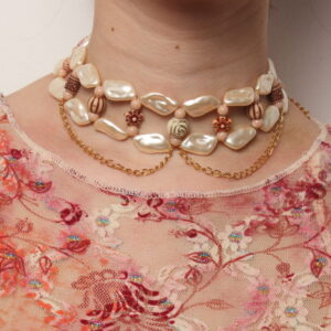 collier tour de cou vintage. Double rangée de perles beiges nacrées en forme de coquillage reliées entre elles par des perles fantaisies fleurs en imitation bois. 2 chaînettes pendantes dorées forment des arcs de cercle. Taille ajustable grâce à la chaînette dorée à l'arrière