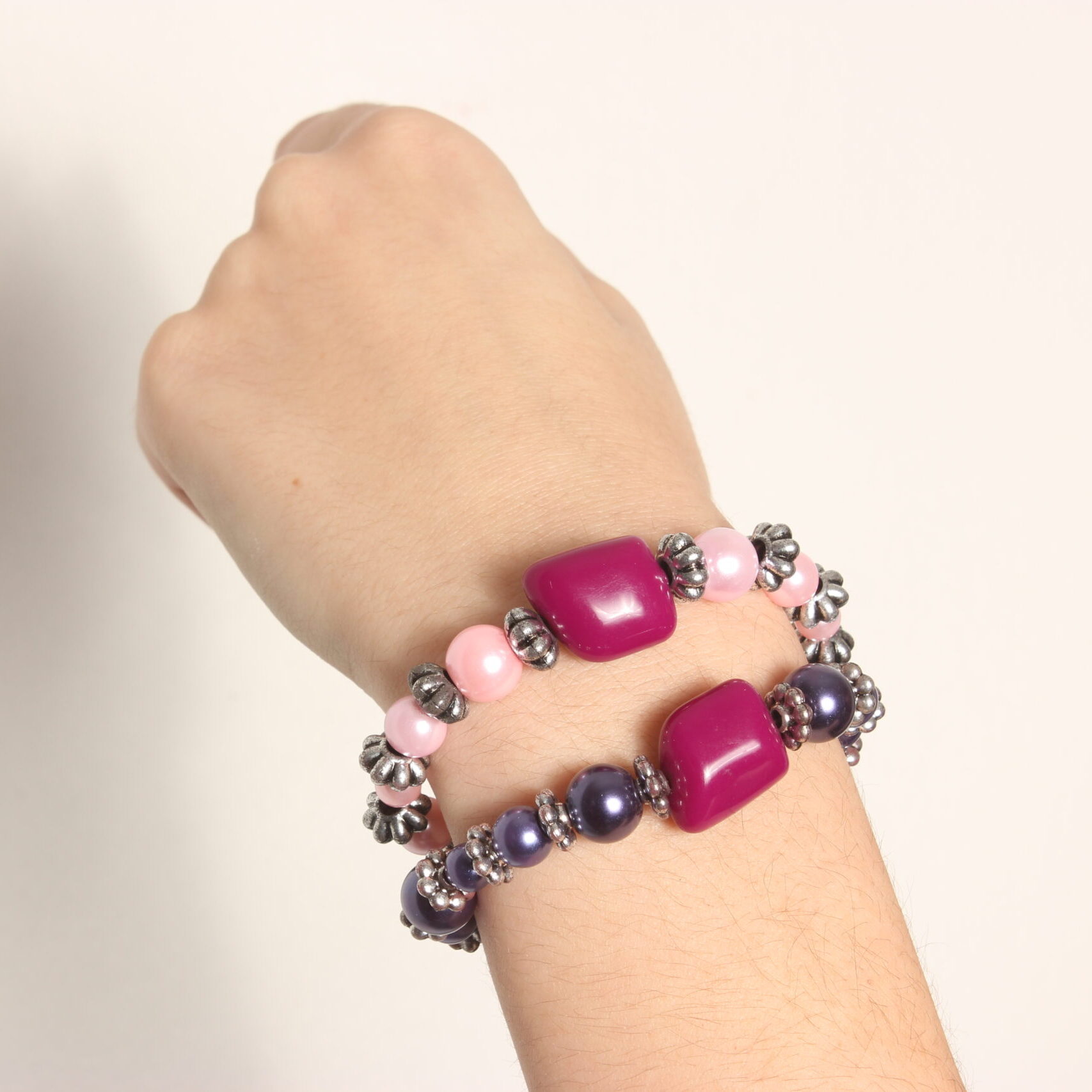 2 bracelets en perles vintage. Un avec une alternance de perles roses pâles et d'intercalaires argentés et l'autre également avec des intercalaires argentés et des perles violettes. Tous 2 rehaussés d'une grosse perle grenat de forme proche du cube.