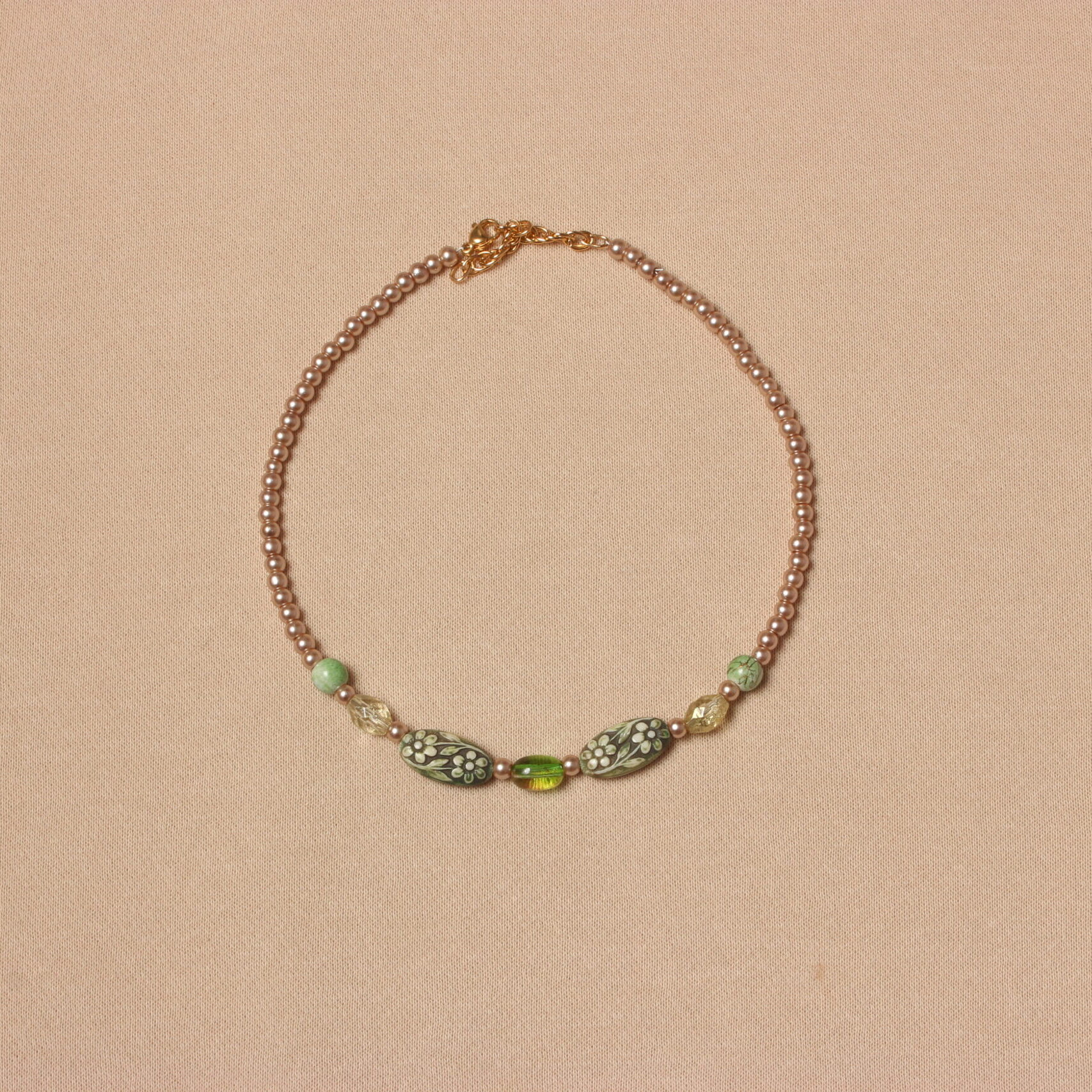 Collier en perles vertes et beiges. Les petites perles beiges se trouvent aux extrémités et sont en verre. Ensuite il y a de chaque côté 2 perles rondes vertes claires striées, puis 2 perles géométriques translucides, 2 perles ovales de taille moyenne, en résine vertes avec des motifs fleurs plus claires et au centre une perle verte translucide. Chaînette d'extension dorée à l'arrière.