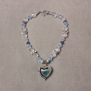 Collier en perles translucides bleues et blanches, et grises argentées. Au centre se trouve un magnifique cœur en verre gris avec un trait noir et un cœur bleu peint au centre. Chaînette d'extension argentée à l'arrière.