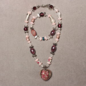 Bracelet et collier en perles. Il y a une alternance de petites perles roses pâles; des moyennes transparentes en forme de goutte, des rondes et des ovales grenat en verre, des grises argentées et des petites rondes transparentes avec des motifs fleurs. Au centre du collier, il y a un pendentif en verre transparent avec un centre rose et des petites fleurs peintes. Chaînette d'extension argentée à l'arrière
