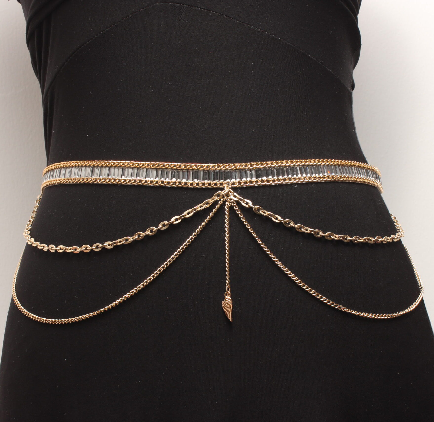 Tour de ventre composé d'une fine ceinture en strass gris enveloppés de 2 chaînettes dorées qui sont plaquées à la ceinture . 2 autres chaînettes dorées (une petite et une plus grande) tombant sur les hanches sont situées de chaque côté. Au centre, il y a une petite chaînette dorée verticale terminée par une petite aile dorée. Chaînette d'extension dorée à l'arrière
