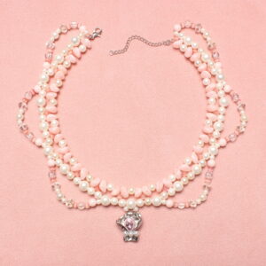 Collier en perles 3 rangs style princesse. La rangées la plus proche du cou est composée d'une alternance de petites perles triangulaires roses pâles et de perles rondes blanches nacrées. La seconde rangée, qui est plaquée contre la première, est composée de perles blanches nacrées. La troisième rangée forme des demis cercles, 2 à droite et 2 à gauche. Elle est composée de perles roses pâles, blanches et translucides. Au centre il y a une breloque argentée agrémentée d'une pierre rose pâle étincelante et de 3 petites perles blanches. Chaînette d'extension argentée à l'arrière.
