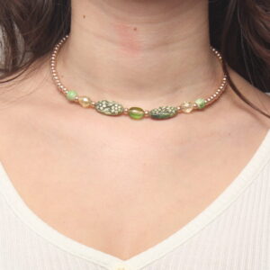 Collier en perles vertes et beiges. Les petites perles beiges se trouvent aux extrémités et sont en verre. Ensuite il y a de chaque côté 2 perles rondes vertes claires striées, puis 2 perles géométriques translucides, 2 perles ovales de taille moyenne, en résine vertes avec des motifs fleurs plus claires et au centre une perle verte translucide. Chaînette d'extension dorée à l'arrière.