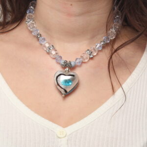 Collier en perles translucides bleues et blanches, et grises argentées. Au centre se trouve un magnifique cœur en verre gris avec un trait noir et un cœur bleu peint au centre. Chaînette d'extension argentée à l'arrière.