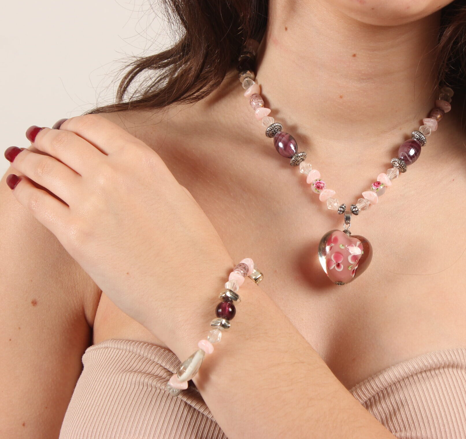 Bracelet et collier en perles. Il y a une alternance de petites perles roses pâles; des moyennes transparentes en forme de goutte, des rondes et des ovales grenat en verre, des grises argentées et des petites rondes transparentes avec des motifs fleurs. Au centre du collier, il y a un pendentif en verre transparent avec un centre rose et des petites fleurs peintes. Chaînette d'extension argentée à l'arrière