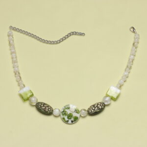 Collier en perles vertes et blanches. Les blanches sont situées aux extrémités. Elles sont en verre. Ensuite se trouvent de chaque côté 2 perles carrées en verre vertes et blanches, 2 perles ovales en résine vertes à motif fleurs et au centre une perle ronde plate en verre blanche avec des feuilles vertes. Chaînette d'extension argentée à l'arrière.