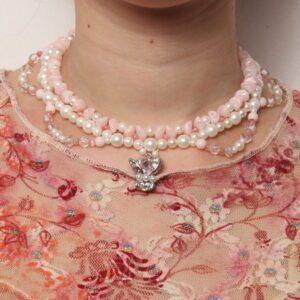 Collier en perles 3 rangs style princesse. La rangées la plus proche du cou est composée d'une alternance de petites perles triangulaires roses pâles et de perles rondes blanches nacrées. La seconde rangée, qui est plaquée contre la première, est composée de perles blanches nacrées. La troisième rangée forme des demis cercles, 2 à droite et 2 à gauche. Elle est composée de perles roses pâles, blanches et translucides. Au centre il y a une breloque argentée agrémentée d'une pierre rose pâle étincelante et de 3 petites perles blanches. Chaînette d'extension argentée à l'arrière.
