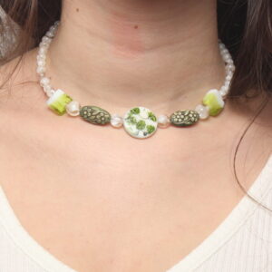 Collier en perles vertes et blanches. Les blanches sont situées aux extrémités. Elles sont en verre. Ensuite se trouvent de chaque côté 2 perles carrées en verre vertes et blanches, 2 perles ovales en résine vertes à motif fleurs et au centre une perle ronde plate en verre blanche avec des feuilles vertes. Chaînette d'extension argentée à l'arrière.