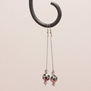 Boucles d'oreilles pendantes. Les apprêts argentés sont en acier inoxydable. Une magnifique perle de céramique est suspendue à une fine chaînette argentée. Des motifs fleurs noirs et gris sont peints sur la perle en céramique blanche. 2 perles à facettes grises claires translucides et de fins intercalaires argentés sont situées au dessus et au dessous de cette perle blanche.