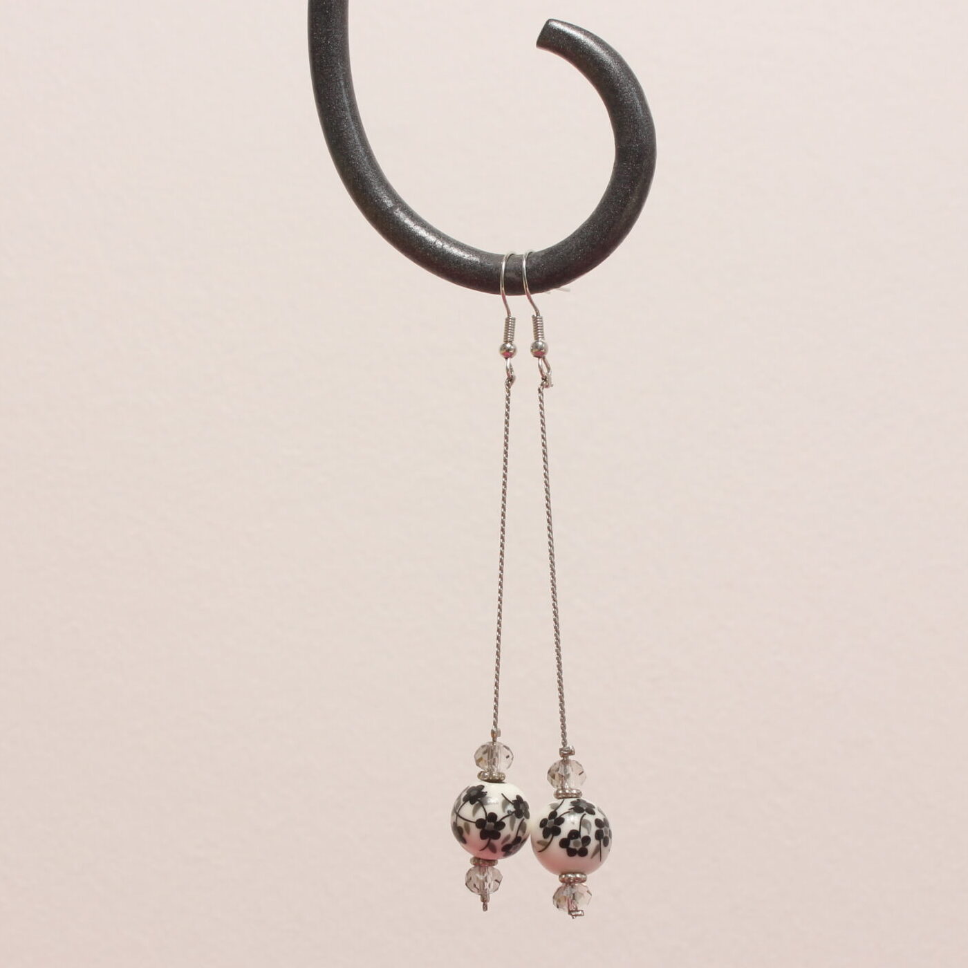 Boucles d'oreilles pendantes. Les apprêts argentés sont en acier inoxydable. Une magnifique perle de céramique est suspendue à une fine chaînette argentée. Des motifs fleurs noirs et gris sont peints sur la perle en céramique blanche. 2 perles à facettes grises claires translucides et de fins intercalaires argentés sont situées au dessus et au dessous de cette perle blanche.