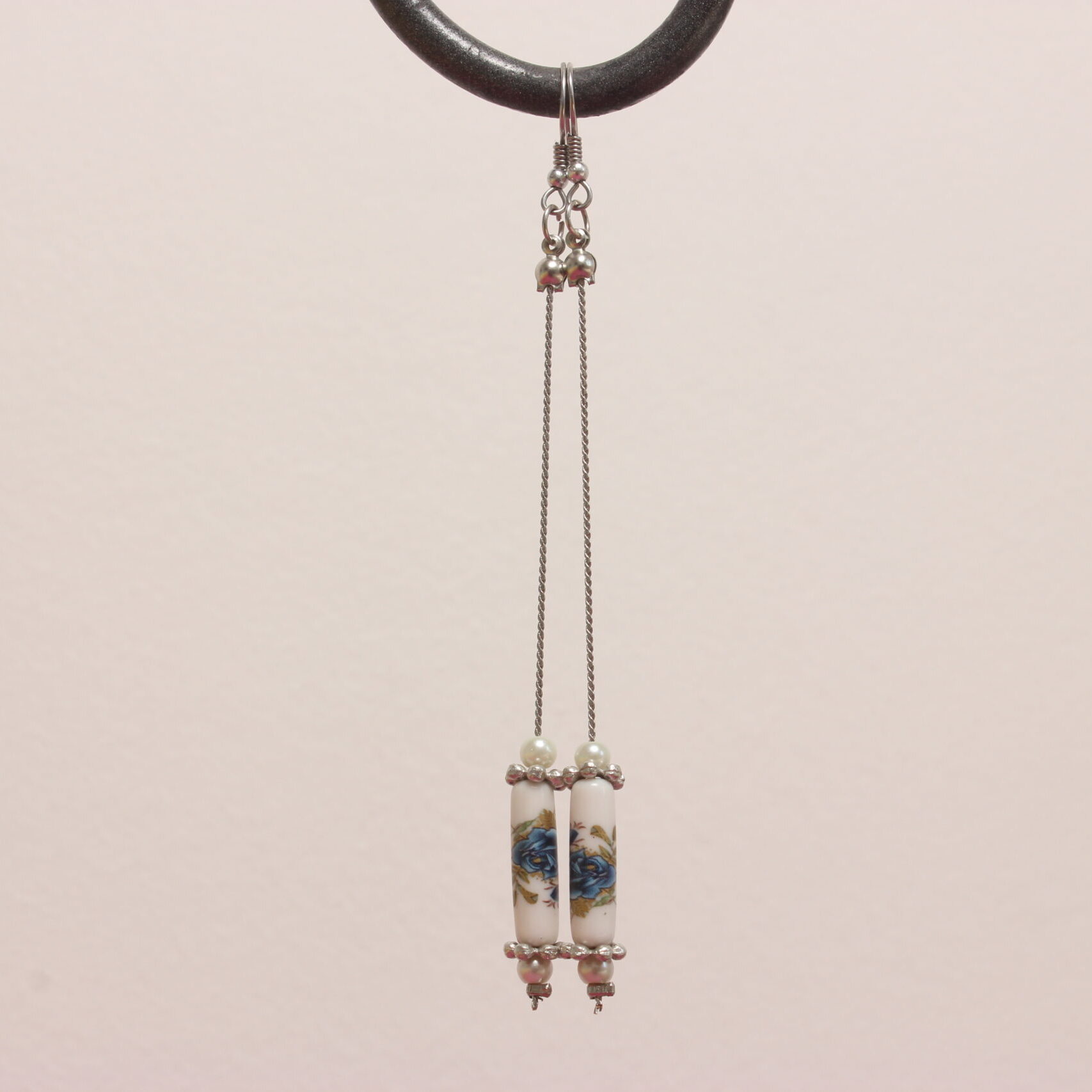 Boucles d'oreilles vintage montées sur des apprêts en acier inoxydable argenté. Magnifique perle en céramique en forme de tube où sont peintes des fleurs bleues au feuillage vert. Elles sont entourées d'intercalaires argentés plats en forme de fleur et de petites perles de verre blanches nacrées.