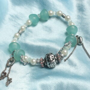 Bracelet en perles. Petites perles blanches nacrées et grises et perles bleues vertes claires translucides en forme de demi cercle qui se font face pour former une sorte de sablier. Au centre une belle breloque grise argentée. De chaque côté 2 breloques en formes de cœur avec des chaînettes argentées tombantes très modernes. Une autre chaînette argentée épouse la forme ronde du bracelet à l'avant au niveau de la breloque centrale ce qui la met en valeur.