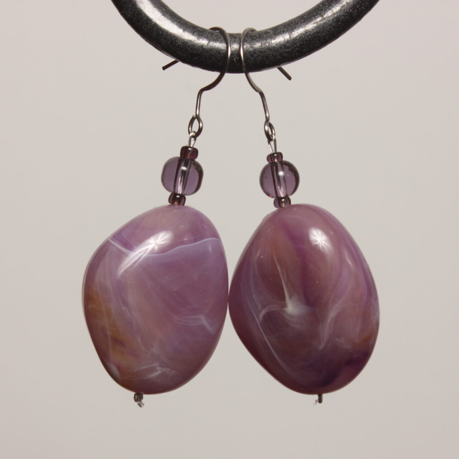 Boucles d'oreilles montées sur des apprêts argentés en acier inoxydable. Grosse perle en forme de galet parme irisée de blanc et ambre. Au-dessus se trouve une perle ronde violette transparente en verre.
