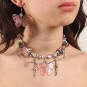 Ensemble collier et boucles d'oreilles. Le collier se compose de perles hexagonales roses pâles et transparentes, rondes blanches en verre, argentées rondes aplaties de tailles variées, transparentes à motif fleur en verre. Au centre il y a un gros pendentif en forme de papillon rose pâle irisé. Des chaînettes argentées tombantes et d'autres en arrondis ornent le devant. Ces chaînettes sont agrémentées de petits cœurs et papillons argentés. Chaînette d'extension argentée à l'arrière. Boucles d'oreilles avec une perle ronde transparente et un pendentif papillon rose pastel comme celui du collier dessous. Apprêts argentés en acier inoxydable