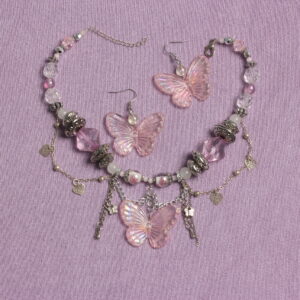 Ensemble collier et boucles d'oreilles. Le collier se compose de perles hexagonales roses pâles et transparentes, rondes blanches en verre, argentées rondes aplaties de tailles variées, transparentes à motif fleur en verre. Au centre il y a un gros pendentif en forme de papillon rose pâle irisé. Des chaînettes argentées tombantes et d'autres en arrondis ornent le devant. Ces chaînettes sont agrémentées de petits cœurs et papillons argentés. Chaînette d'extension argentée à l'arrière. Boucles d'oreilles avec une perle ronde transparente et un pendentif papillon rose pastel comme celui du collier dessous. Apprêts argentés en acier inoxydable