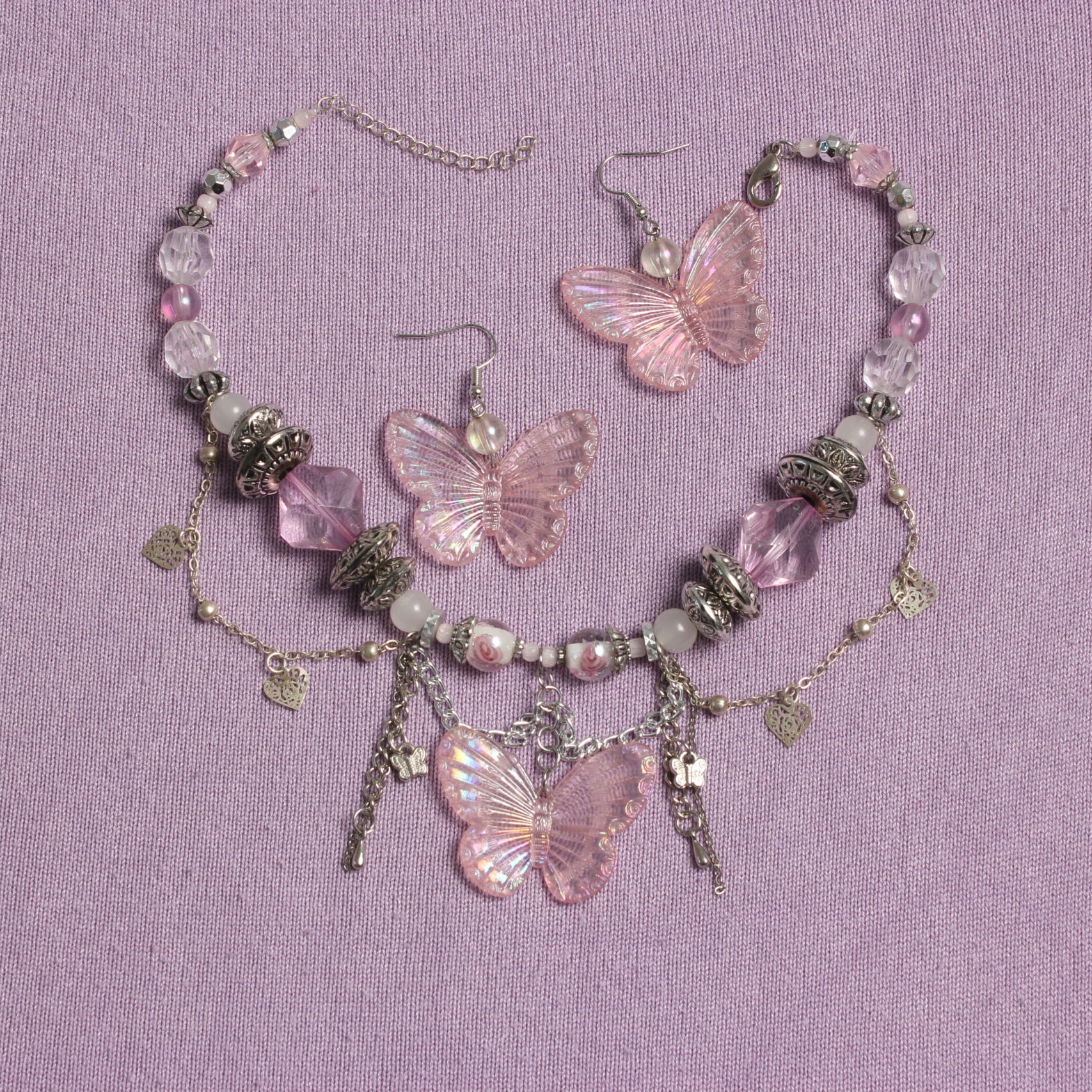 Ensemble collier et boucles d'oreilles. Le collier se compose de perles hexagonales roses pâles et transparentes, rondes blanches en verre, argentées rondes aplaties de tailles variées, transparentes à motif fleur en verre. Au centre il y a un gros pendentif en forme de papillon rose pâle irisé. Des chaînettes argentées tombantes et d'autres en arrondis ornent le devant. Ces chaînettes sont agrémentées de petits cœurs et papillons argentés. Chaînette d'extension argentée à l'arrière. Boucles d'oreilles avec une perle ronde transparente et un pendentif papillon rose pastel comme celui du collier dessous. Apprêts argentés en acier inoxydable