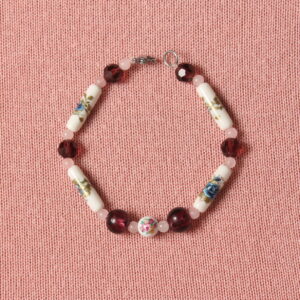 Bracelet composé d'une alternance de petites perles roses pâles en verre, des moyennes hexagonales prunes, des longues en forme de tube en céramique blanche à motif fleur bleue et verte. Au centre se trouve une perle ronde en céramique blanche avec une fleur rose entourée de perles rondes en verre prunes. Fermoir argenté