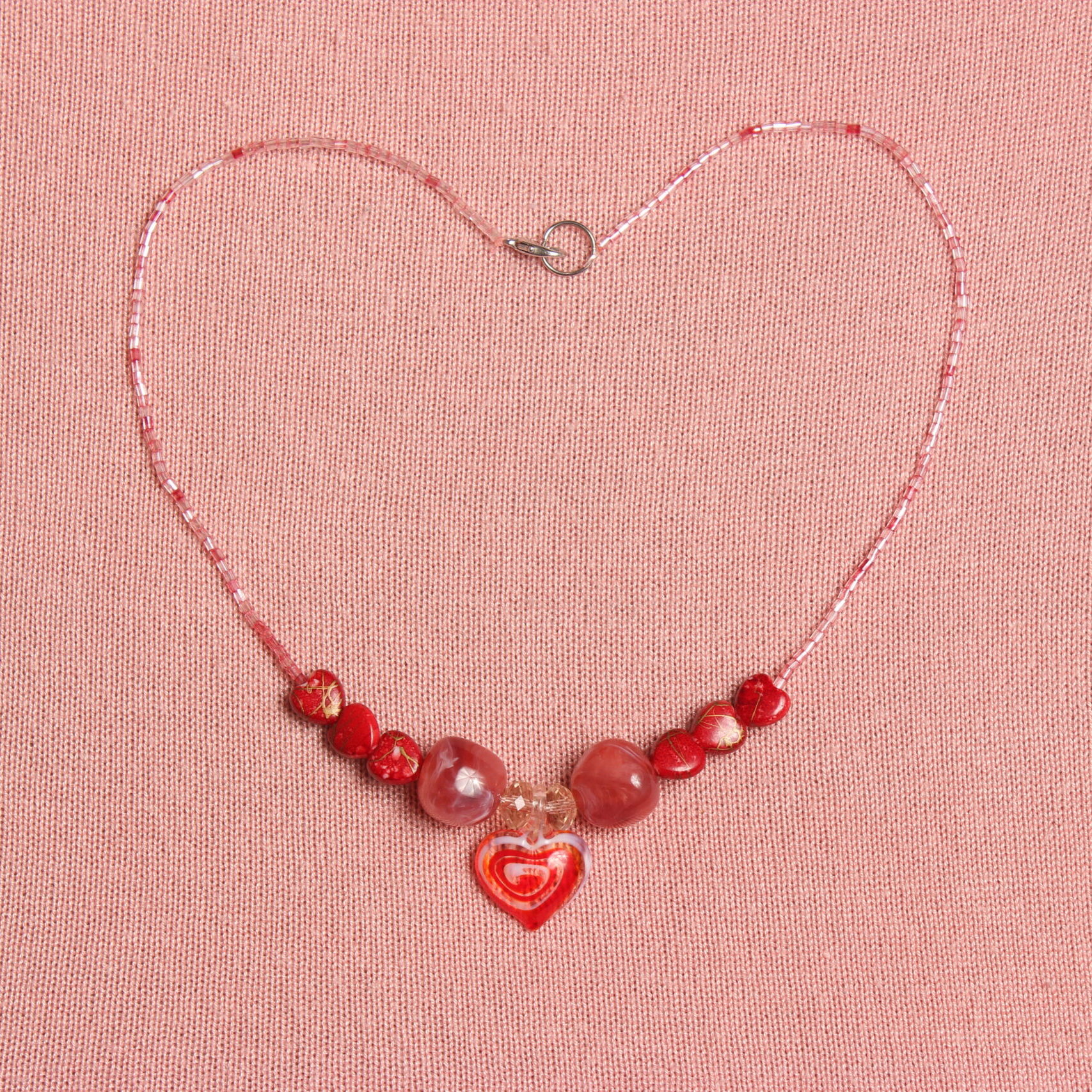 Collier composé de chaque côté de 3 petites perles en forme de cœur, 1 grosse perle rouge rosée, 1 perle en verre transparente et au centre un pendentif en verre rouge et blanc en forme de cœur.