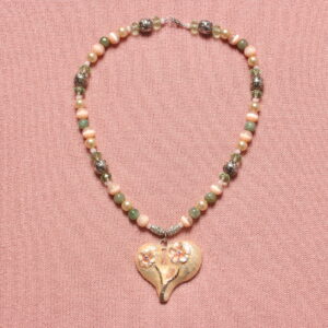Collier style vintage alternant les perles rondes de taille moyenne roses crevettes, vertes, beiges, des petites perles beiges, des perles hexagonales vertes translucides et des grosses perles argentées à motif tribal. Au centre se trouve un gros pendentif en forme de cœur rose crevette où se trouvent 2 fleurs aux cœurs formés de strass roses étincelants. Fermoir argenté