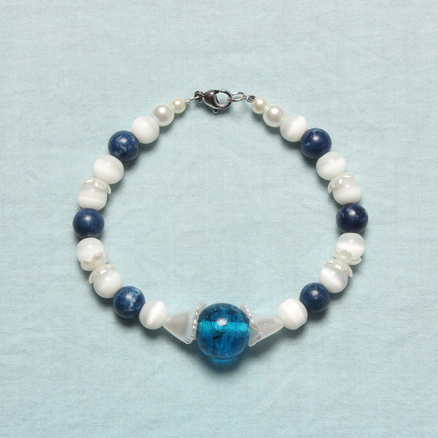 Bracelet composé de perles en pierre blanches et bleues marines upcyclées. De petites fleurs blanches nacrées apportent une délicate touche printanière. Il est rehaussé d’une magnifique perle upcyclée bleue roi en verre au centre enveloppé de 2 fleurs blanches nacrées.