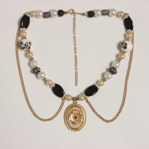 Ce collier se compose de perles rondes blanches moyennes, beiges petites, blanches grosses en céramique avec des fleurs noires et grises peintes et qui sont entourées d'intercalaires dorés. Il y a aussi des perles ovales plates noires entourées de 2 corolles blanches nacrées. 2 perles de lave noire se trouvent aux extrémités du collier. Il y a aussi des perles en résine beige en forme de tonneau et des grises rondes aplaties. 2 chaînes dorées pendent sur le torse. Au centre se trouve un médaillon ovale doré avec une fleur dorée en relief et une perle scintillante rouge au cœur de la fleur. Chaînette d'extension dorée à l'arrière