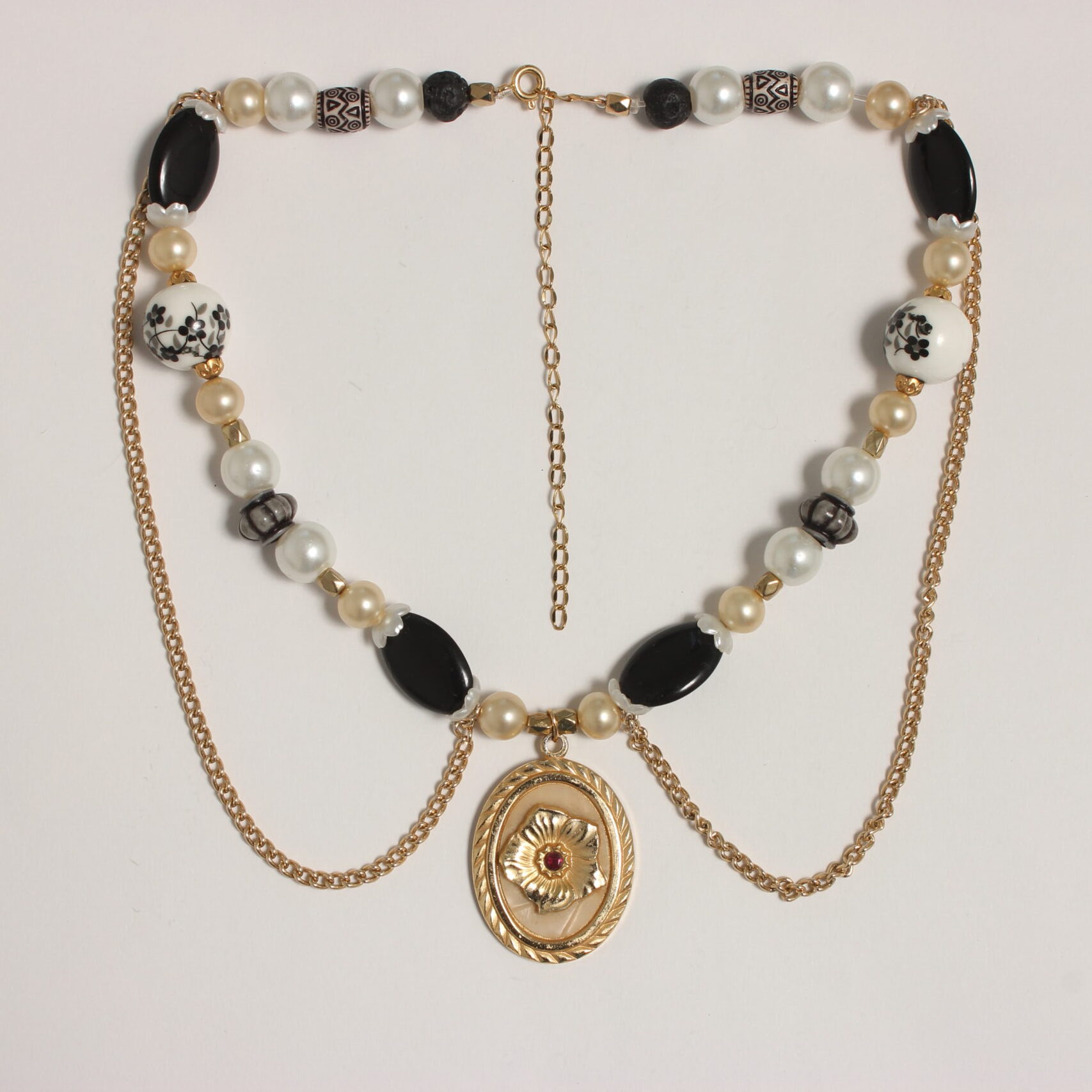 Ce collier se compose de perles rondes blanches moyennes, beiges petites, blanches grosses en céramique avec des fleurs noires et grises peintes et qui sont entourées d'intercalaires dorés. Il y a aussi des perles ovales plates noires entourées de 2 corolles blanches nacrées. 2 perles de lave noire se trouvent aux extrémités du collier. Il y a aussi des perles en résine beige en forme de tonneau et des grises rondes aplaties. 2 chaînes dorées pendent sur le torse. Au centre se trouve un médaillon ovale doré avec une fleur dorée en relief et une perle scintillante rouge au cœur de la fleur. Chaînette d'extension dorée à l'arrière