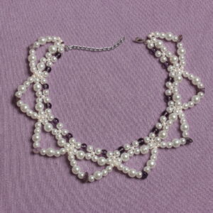 ce collier entièrement en verre est constitué de trois rangées. Deux très rapprochées en ras du cou et une troisième plus grande formant des demi cercles. Ce collier possède une alternance de perles nacrées blanches et de perles violettes. Ainsi qu'une autre alternance entre des perles nacrées blanches plus ou moins grosses. Ce bijou se ferme grâce à un petit fermoir et à une petite chaînette.