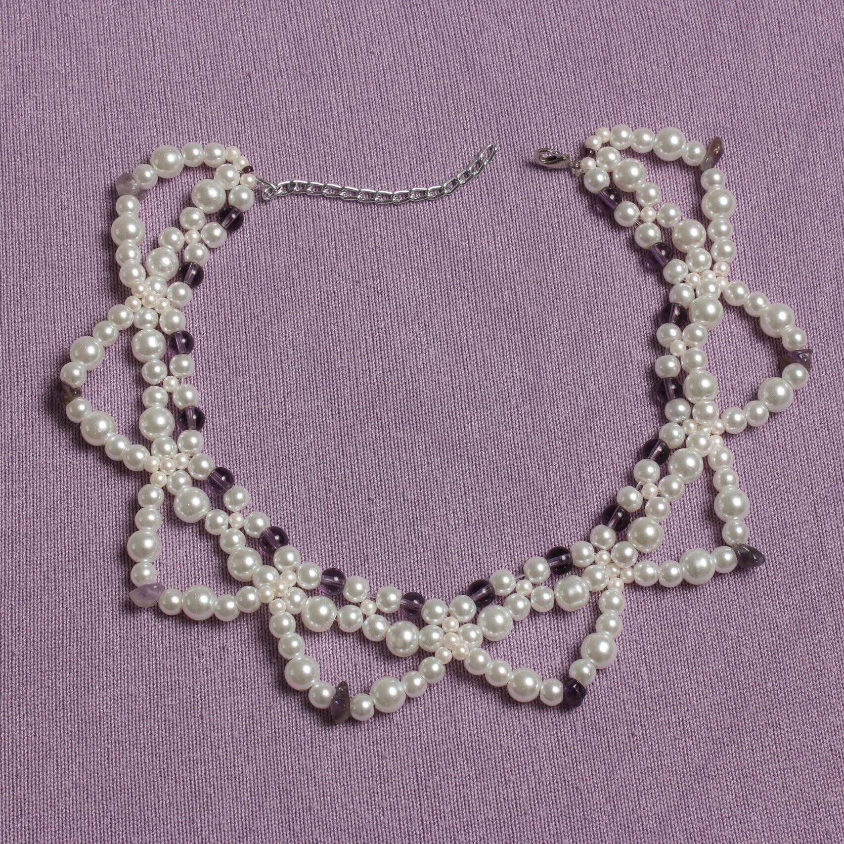 ce collier entièrement en verre est constitué de trois rangées. Deux très rapprochées en ras du cou et une troisième plus grande formant des demi cercles. Ce collier possède une alternance de perles nacrées blanches et de perles violettes. Ainsi qu'une autre alternance entre des perles nacrées blanches plus ou moins grosses. Ce bijou se ferme grâce à un petit fermoir et à une petite chaînette.