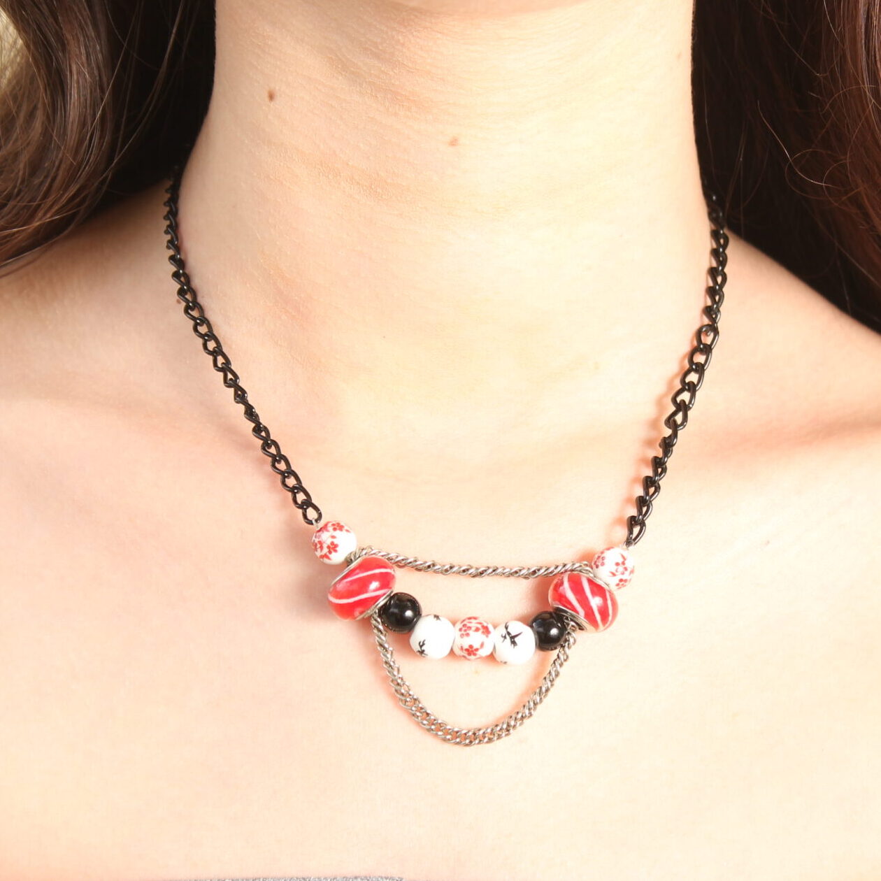 Collier d'inspiration japonaise. Il est composé d'une chaîne noire et d'une série de perles au centre: 2 perles en céramique blanches au motifs japonais rouges, suivies de 2 perles en verre rouges striées de blanc, un peu plus grosses et au bords aplatis, puis 2 perles noires unies, 2 perles de céramique blanches à motifs japonais noirs et une dernière au milieu blanche à motifs rouges identique aux premières. Une chaînette argentée se trouve au dessus des perles rouges et une autre chaînette pend dessous les perles rouges en les reliant.