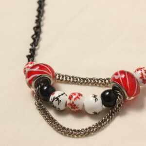 Collier d'inspiration japonaise. Il est composé d'une chaîne noire et d'une série de perles au centre: 2 perles en céramique blanches au motifs japonais rouges, suivies de 2 perles en verre rouges striées de blanc, un peu plus grosses et au bords aplatis, puis 2 perles noires unies, 2 perles de céramique blanches à motifs japonais noirs et une dernière au milieu blanche à motifs rouges identique aux premières. Une chaînette argentée se trouve au dessus des perles rouges et une autre chaînette pend dessous les perles rouges en les reliant.