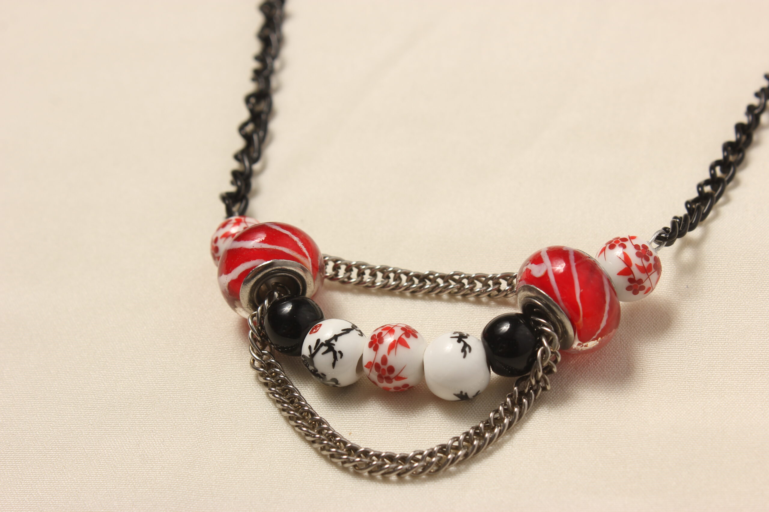 Collier d'inspiration japonaise. Il est composé d'une chaîne noire et d'une série de perles au centre: 2 perles en céramique blanches au motifs japonais rouges, suivies de 2 perles en verre rouges striées de blanc, un peu plus grosses et au bords aplatis, puis 2 perles noires unies, 2 perles de céramique blanches à motifs japonais noirs et une dernière au milieu blanche à motifs rouges identique aux premières. Une chaînette argentée se trouve au dessus des perles rouges et une autre chaînette pend dessous les perles rouges en les reliant.