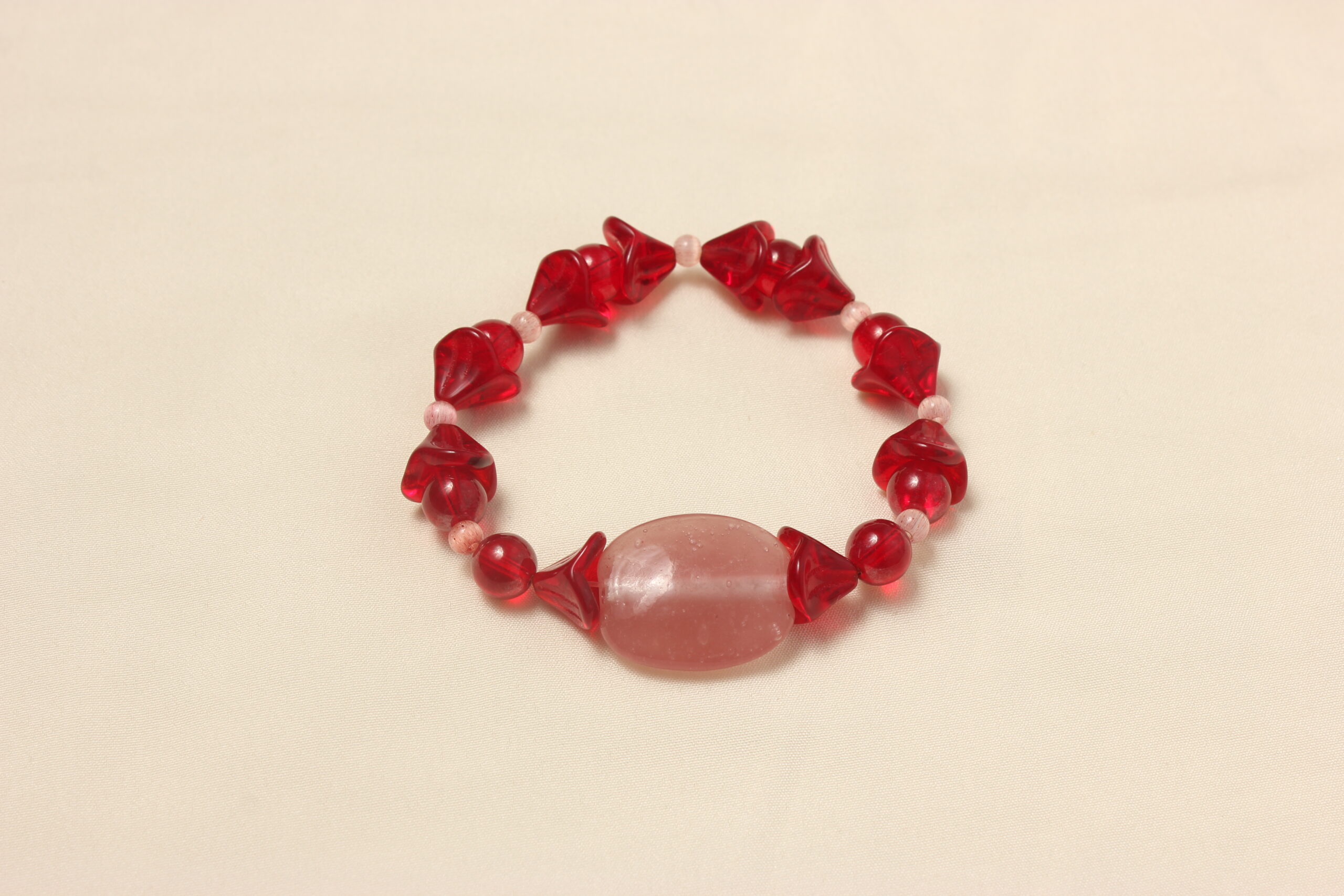 Ce bracelet est constitué d'une grosse perle ovale et rose au centre. symétriquement à cette perle il y a une alternance entre des perles rouges rondes, des perles rouges en formes de fleur et d'autres plus petites œil de chat roses.