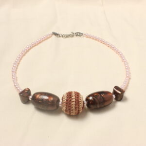 Ce collier est disposé symétriquement autour d'une perle en bois qui est recouverte d'une couverture en crochet marron et beige. autour est disposé une longue perle marron avec des détails marbrés et une autre perle dans les mêmes tons plus petite. chaque perles sont séparées par des petites perles nacrées rose. Le collier se fini par une suite de petites perles roses nacrées. Il est ensuite fermé par une chaînette et un fermoir.