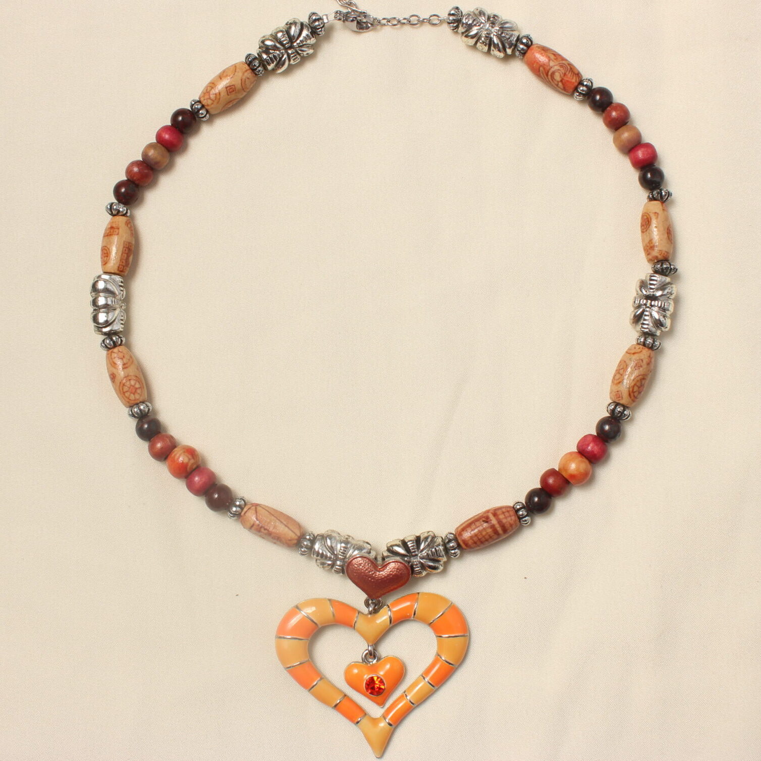 Ce collier est constitué d'un gros pendentif en forme de cœur rayé orange et rouge. au dessus de ce cœur est disposé un deuxième plus petit et orange également. A l’intérieur du gros cœur se trouve encore un petit orange avec une strass rouge au centre. Ce collier est symétrique autour du gros pendentif. il y a d'abord une perle argentée ovale avec des petits détails, suivie d'une perle allongée en bois avec des petits détails marrons et rouges entourées d'intercalaires argentées. ensuite il y a une suite de perles disposées symétriquement autour d'une perle en bois orange. Une petite perle en pierre marron, et une autre en bois rouge. Ensuite. il y a deux perles longues en bois entourées de petites intercalaires qui sont autour d'une perle longue argentée. l'alternance de perles en bois et en pierre recommence avant de finir par une longue perle en bois entourée d'intercalaires et suivie d'une dernière perle argentée. Le collier se finit avec une petite chaînette d’extension ainsi qu'un fermoir pour fermer le collier.