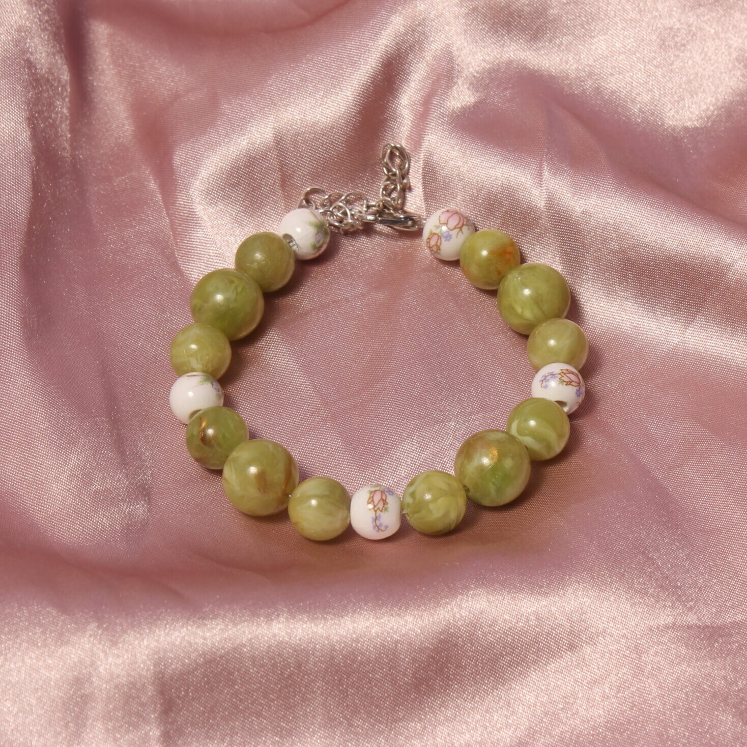Ce bracelet est constitué de 5 perles en céramique blanches et roses avec des petits détails fleuri. entre elles se trouve des perles vertes de différentes tailles.