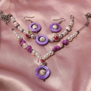 Cette parure est composée d’un bracelet, d’un collier et d’une paire de boucles d’oreilles. Les trois bijoux possèdent au centre une perle nacrée violette, ronde et plate, ornée en son centre d’une petite perle en verre ainsi que d’une perle en céramique rose et blanche aux délicats motifs fleuris. Le bracelet est composé d’une alternance de perles nacrées blanches en verre ciré, de perles en céramique roses, ainsi que de deux perles en céramique plus imposantes, toujours ornées de motifs fleuris. Le collier reprend cette même harmonie avec une alternance de perles nacrées blanches, de petites perles en céramique et de perles en céramique plus imposantes. Il est également agrémenté de petites perles plates violettes. Une chaîne dorée se prolonge ensuite jusqu’aux extrémités du collier, qui se ferme à l’aide d’une petite chaînette et d’un fermoir, comme le bracelet.