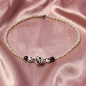 Ce collier possède en son centre une grosse perle noire et blanche en céramique avec des motifs floraux. autour de celle ci sont mises symétriquement une perle en verre allongée, une petite perle en céramique noire et blanche avec des petits détails et une perle noire. Des perles blanches en verre cirée vont jusqu'à la fin du collier.