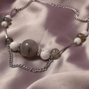 Ce collier est construit autour d’une grosse perle centrale grise. Autour de celle-ci sont disposés des intercalaires, suivis de perles blanches et grises. Il y a ensuite une longue perle grise qui sépare cette partie d’une autre, constituée de deux perles blanches autour d’une perle grise. Une autre longue perle est placée avant de terminer le collier par une chaînette. Celui-ci se ferme grâce à un fermoir. Il possède également une chaînette au centre qui tombe légèrement.
