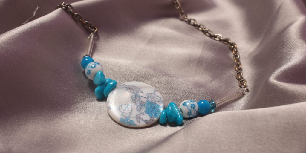 Ce collier est constitué d'une perle centrale ronde et plate en céramique avec des petits détails en forme de feuilles et de grappe de raisin. Autour de celle ci sont mises symétriquement trois jolies petites perles plates, elles sont suivies d'une autre perle en céramique bleue et blanche ronde avec pour motifs des petites fleurs. Il y a ensuite une petite perle ronde bleue et enfin une perle en verre bleue suivie d'une longue perle en verre transparente. le collier se termine avec une longue chaîne et un fermoir pour pouvoir le fermer.