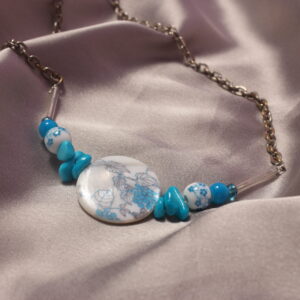 Ce collier est constitué d'une perle centrale ronde et plate en céramique avec des petits détails en forme de feuilles et de grappe de raisin. Autour de celle ci sont mises symétriquement trois jolies petites perles plates, elles sont suivies d'une autre perle en céramique bleue et blanche ronde avec pour motifs des petites fleurs. Il y a ensuite une petite perle ronde bleue et enfin une perle en verre bleue suivie d'une longue perle en verre transparente. le collier se termine avec une longue chaîne et un fermoir pour pouvoir le fermer.