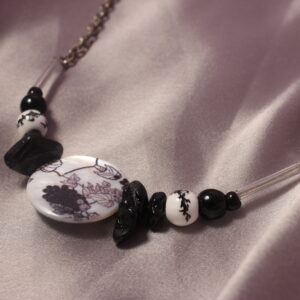 Ce collier est constitué d'une perle centrale ronde et plate en céramique avec des petits détails en forme de feuilles et de grappe de raisin. Autour de celle ci sont mises symétriquement trois jolies petites perles plates, elles sont suivies d'une autre perle en céramique noires et blanche ronde avec des motifs. Il y a ensuite une petite perle ronde noir et enfin une perle en verre noire suivie d'une longue perle en verre transparente. le collier se termine avec une longue chaîne et un fermoir pour pouvoir le fermer.
