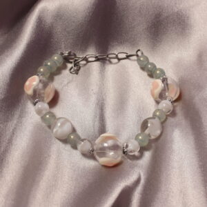 Ce bracelet est composé de perles en verre. Il possède trois perles transparentes ornées de détails roses et blancs, formant un motif en cœur. Entre chacune de ces perles se trouvent de plus petites perles grises ou blanches, apportant une touche délicate à l’ensemble. Le bracelet se ferme à l’aide d’un fermoir et d’une petite chaînette, permettant également d’ajuster légèrement sa longueur.