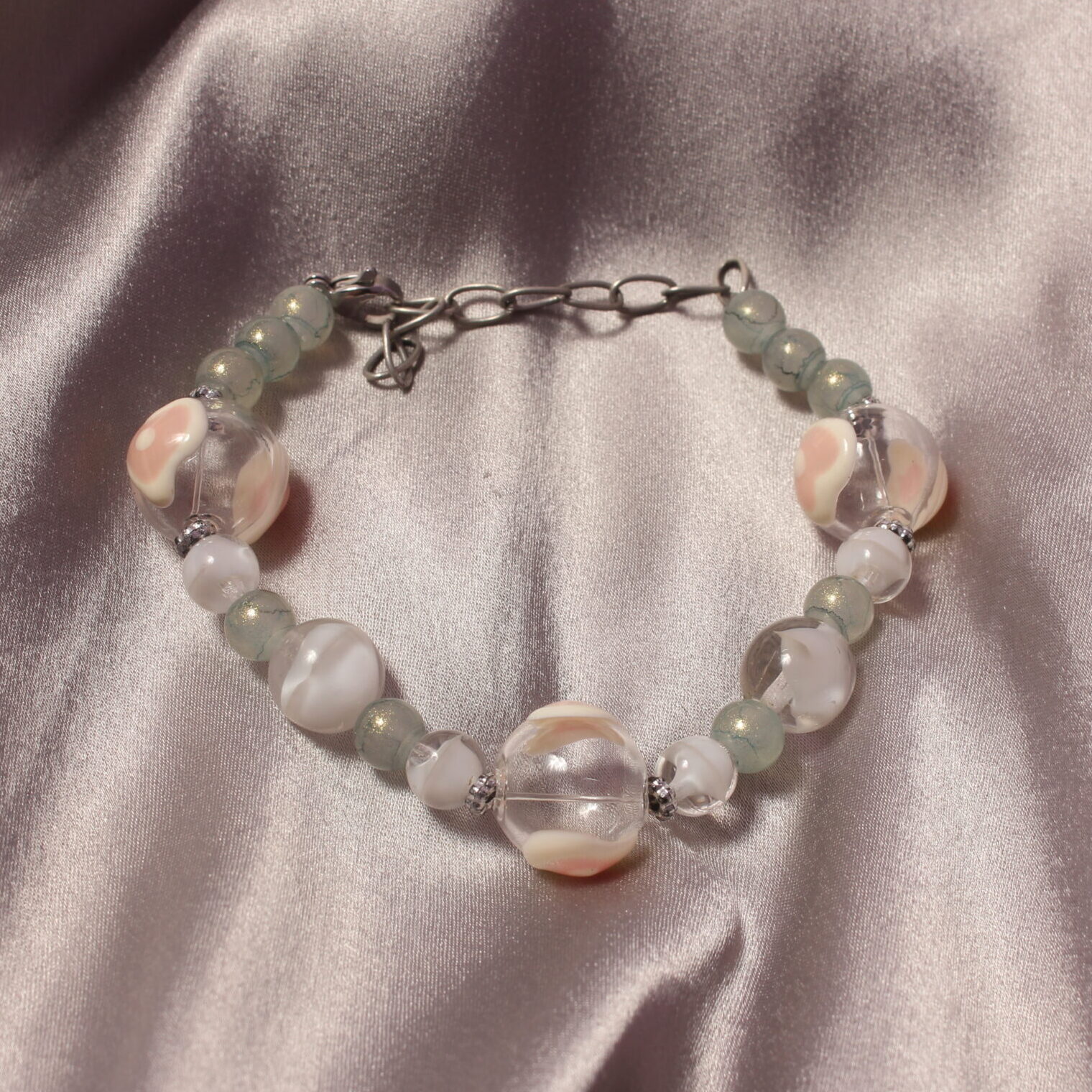 Ce bracelet est composé de perles en verre. Il possède trois perles transparentes ornées de détails roses et blancs, formant un motif en cœur. Entre chacune de ces perles se trouvent de plus petites perles grises ou blanches, apportant une touche délicate à l’ensemble. Le bracelet se ferme à l’aide d’un fermoir et d’une petite chaînette, permettant également d’ajuster légèrement sa longueur.