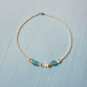 Ce collier plutôt fin est construit de manière symétrique autour d’une petite perle blanche, qui semble être en pierre. Autour de celle-ci se trouvent deux perles en céramique jaune et blanche, ornées d’un motif de tournesol. Deux perles bleues en verre, de forme allongée, sont ensuite disposées de chaque côté. Elles sont suivies d’une autre perle en céramique ornée d’un motif de tournesol. Le collier se poursuit ensuite avec une succession de perles blanches nacrées en verre ciré jusqu’à son extrémité. Il se ferme grâce à un anneau et à un petit fermoir.