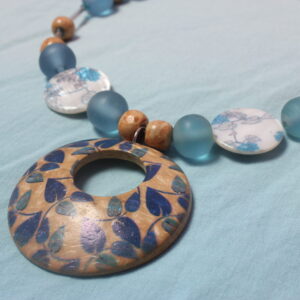 Ce collier est constitué d'un pendentif central en bois avec des détails en formes de feuilles bleues. Autour de celle ci sont installées des perles de différentes tailles et formes bleu et marron, en bois, en verre et en céramique.