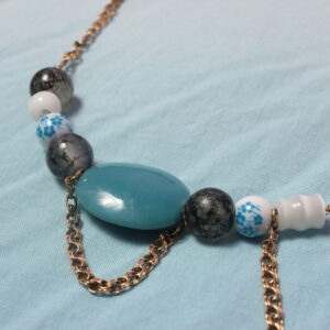 Ce magnifique collier asymétrique est construit autour d’une grosse perle ovale bleue, probablement en pierre ou en céramique. Elle est délicatement encadrée par une chaînette dorée. Sur son côté droit se trouvent une perle grise, une perle blanche et bleue en céramique ornée d’un motif fleuri, une perle blanche probablement en pierre, ainsi qu’une autre perle grise. De l’autre côté, on retrouve une perle grise, une perle bleue en céramique au motif fleuri, ainsi qu’une perle blanche plus allongée, là encore probablement en pierre. Entre ces deux dernières perles se trouve une petite chaînette dorée qui tombe délicatement, apportant une légère touche de mouvement au collier. Le collier se poursuit avec une chaîne dorée jusqu’à ses extrémités et se ferme à l’aide d’un anneau et d’un fermoir doré.